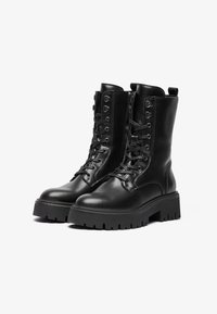 Bottes de combat en cuir noir avec une tige haute, bout rond, lacets à l'avant, fermeture éclair sur le côté et une semelle en caoutchouc chunky avec un motif rainuré.