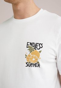 Fehér pamut póló, amelyen egy sárga-narancs citrusgyümölcs és zöld levelek grafikája látható, és a "VÉGTELEN NYÁR" felirat szerepel felül és alul.