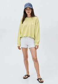 Felpa cropped verde lime con un piccolo logo, abbinata a pantaloni corti in denim bianco strappati e infradito nere, completata da un cappellino blu.