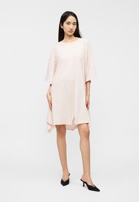 Ανοιχτό ροζ φαρδιά φόρεμα με κοντά μανίκια, με άνετη εφαρμογή και ασύμμετρο τελείωμα. Φοράται με μαύρες ψηλοτάκουνες γόβες με μυτερή μύτη.