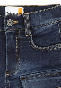 Jeans in denim blu scuro con cuciture gialle, dotati di un bottone metallico, tasche frontali e un'etichetta con il logo Timberland sulla vita.