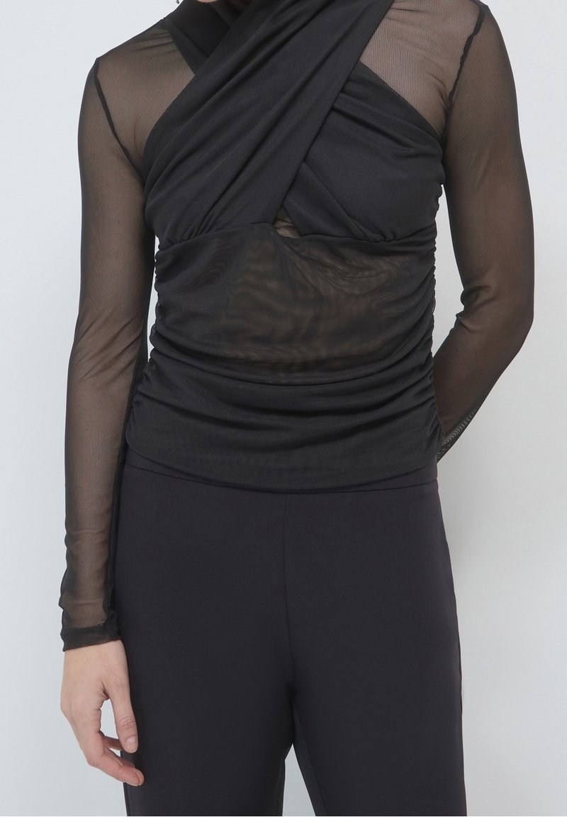 Femme portant un haut noir transparent à manches longues avec un détail croisé sur un sous-vêtement noir et un pantalon noir, debout devant un fond uni.