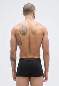 Černé boxerky s vínovým pasem, zdobené logem značky. Model má na zádech viditelné tetování a krátké vlasy.