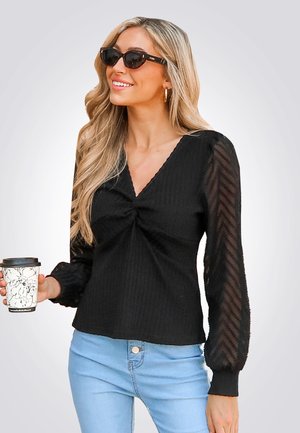 Top negro de canalé con escote en V y detalle frontal retorcido. Mangas largas semitransparentes con patrón de chevron. Combinado con jeans azul claro y sosteniendo una taza de café.