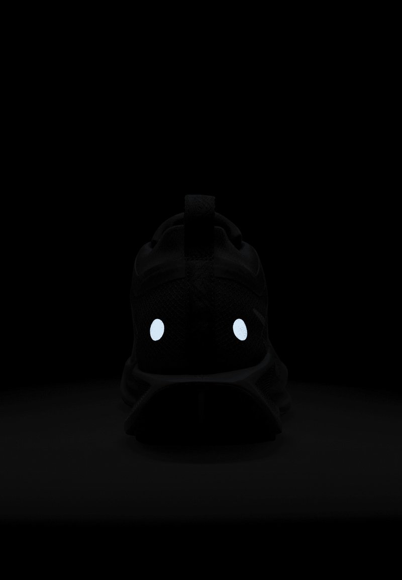 Zapatilla negra con detalles reflectantes en forma de ojos, parte superior de malla texturizada y una etiqueta de tirón en el talón, presentada sobre un fondo oscuro.