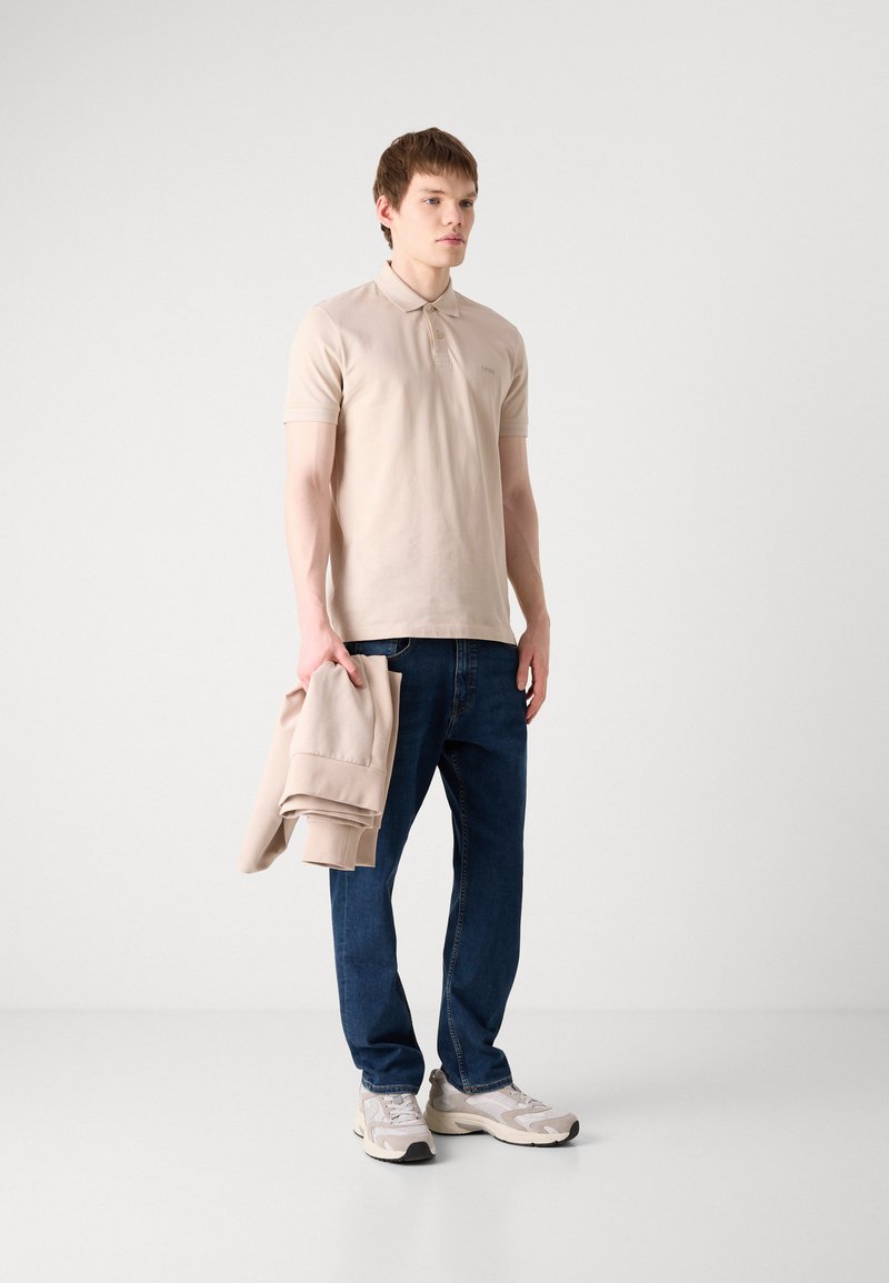 Polo beige avec un col, associé à un jean bleu foncé. Le mannequin tient un sweat-shirt beige, portant des baskets claires. Design simple, tissu doux.