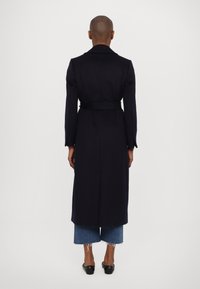 Tmavomodrý trenčkot s páskem v pase, dlouhými rukávy a na míru šitým střihem, kombinovaný s oříznutými džínovými kalhotami a černými balerínami.