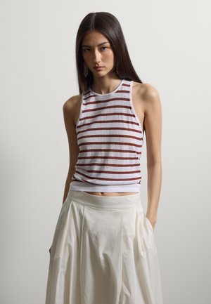 Femme aux longs cheveux bruns portant des boucles d’oreilles en or, un haut blanc sans manches avec des rayures horizontales rouges, et une jupe blanche taille haute.