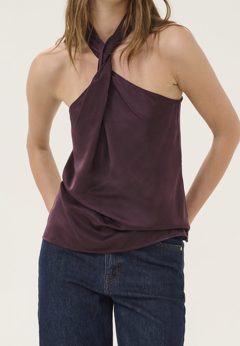 Femme portant un haut sans manches violet foncé col halter associé à un jean bleu foncé sur un fond clair uni.