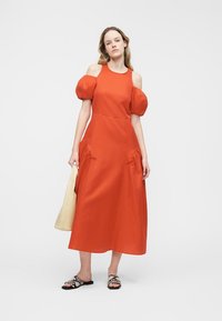 Γυναίκα που φοράει midi φόρεμα κόκκινου χρώματος με σκάλες στους ώμους και φουσκωτά μανίκια, κρατάει μπεζ τσάντα και φοράει μαύρα και άσπρα σανδάλια, στέκεται σε λευκό φόντο.