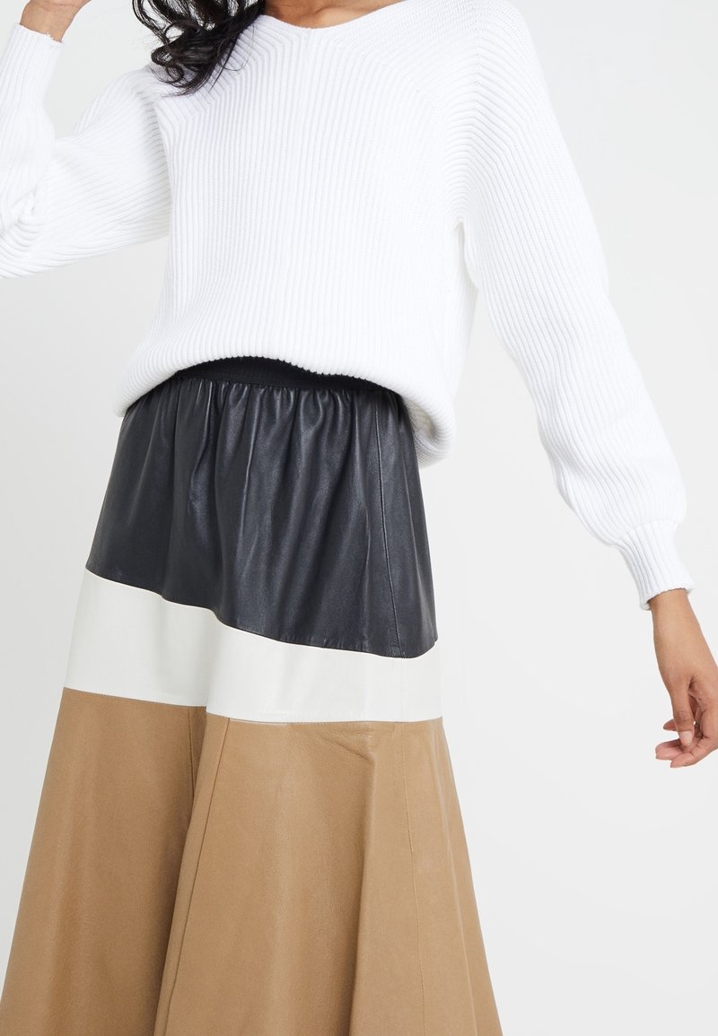 Femme portant un pull blanc côtelé et une jupe taille haute à blocs de couleurs avec des panneaux de cuir noirs, blancs et beige sur un fond uni.