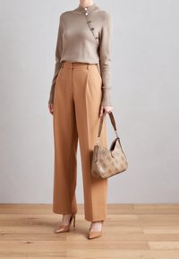 Beige Strickpullover mit hohem Kragen und seitlichen Knopfdetails, kombiniert mit hochgeschnittenen, weiten, hellbraunen Hosen und einer gemusterten Handtasche.
