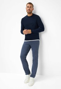 Maritimer Strickpullover mit Rundhalsausschnitt, kombiniert mit dunkelblauen Slim-Fit-Chinos. Die Schuhe sind weiße Ledersneaker. Vor einem einfarbigen Hintergrund.