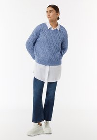 Lichtblauwe gebreide trui met een textuurpatroon, gedragen over een witte blouse met kraag, gecombineerd met donkerblauwe spijkerbroek en witte sneakers.