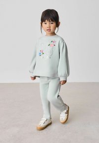 Jasnozielony sweatshirt z kwiatowym nadrukiem, w połączeniu z pasującymi spodniami z fakturowanymi akcentami oraz białymi butami z podwójnymi klamrami.