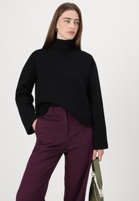Svart strikkegenser med høy hals og vide ermer, kombinert med dype plommefargede skreddersydde bukser. Modellen holder en grønn håndveske.