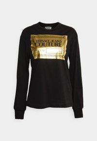 Camisa de manga larga negra con impresión de logo metálico dorado que dice "Versace Jeans Couture" y un borde decorativo. Textura suave. Cuello redondo.