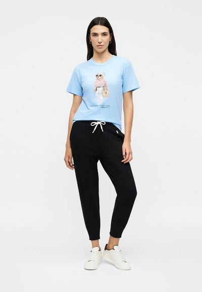 Γυναίκα που φοράει ένα ανοιχτό μπλε t-shirt με σχέδιο αρκούδας, μαύρο αθλητικό παντελόνι με λευκό κορδόνι και λευκά αθλητικά παπούτσια, στέκεται μπροστά σε απλό φόντο.