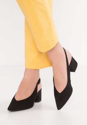 Sorte ruskinds slingback hæle med spids tå og en chunky hæl. Har en V-formet udskæring og justerbar rem. Båret sammen med gule cropped bukser.