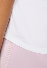 Bijela sportska majica s DRYCELL tehnologijom koja odvodi vlagu, uparen s laganim ružičastim tajicama koje imaju teksturirani uzorak i glatku završnicu.