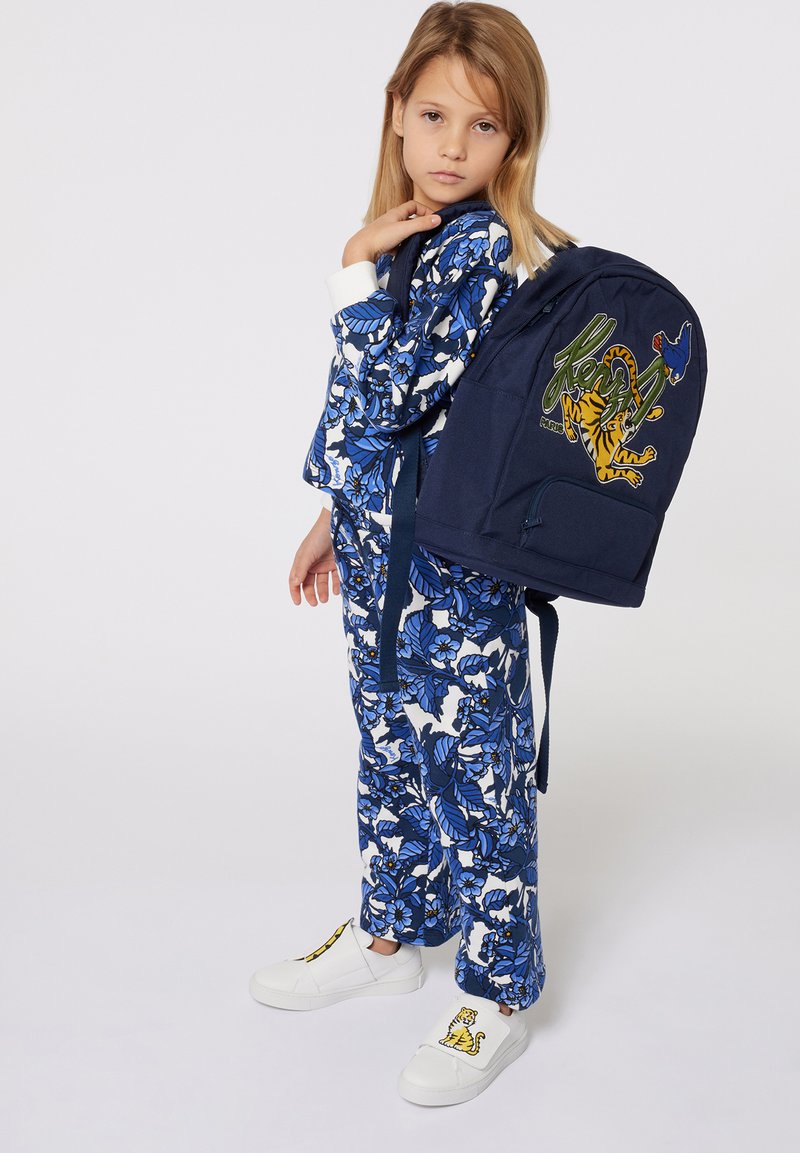 Sac à dos marine avec des patches de tigre et d'oiseau, tenue bleue et blanche à motif floral, et chaussures blanches avec un design de tigre.