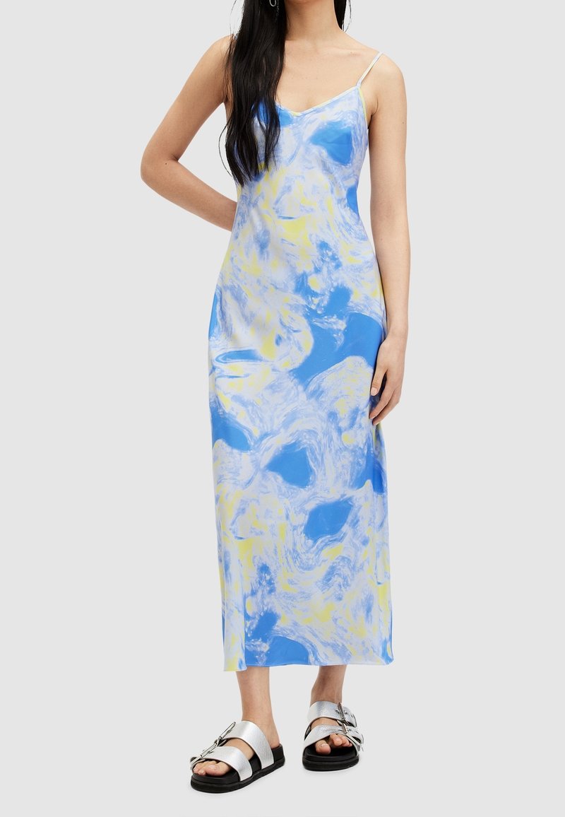 Femme portant une robe midi sans manches marbrée bleue et jaune avec de fines bretelles et des sandales à boucle argentée, debout avec un bras plié.