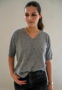 Jeune femme aux cheveux bruns en queue de cheval portant un pull gris à manches courtes, un jean noir, une montre dorée, des bagues et des colliers à plusieurs couches, regardant vers la gauche.