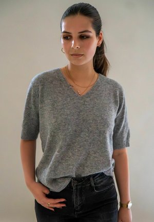 Jonge vrouw met donker haar in een paardenstaart, gekleed in een grijze, kort aangesloten trui, zwarte jeans, een gouden horloge, ringen en gelaagde kettingen, die naar links kijkt.