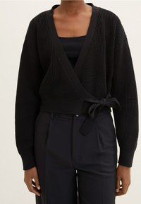 Černý pletený cardigan s hlubokým V výstřihem, širokými rukávy a vázáním v pase, kombinovaný s černým tílkem a tmavými kalhotami.