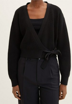 Czarny dziany kardigan w kształcie kopertowym z głębokim dekoltem w serek, szerokimi rękawami i wiązaniem w talii, w zestawieniu z czarnym topem na ramiączkach i ciemnymi spodniami.