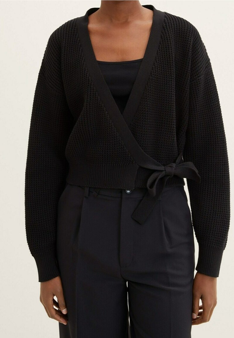 Černý pletený cardigan s hlubokým V výstřihem, širokými rukávy a vázáním v pase, kombinovaný s černým tílkem a tmavými kalhotami.