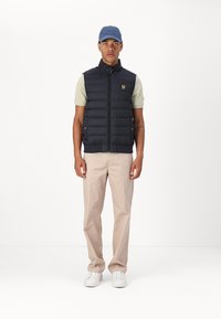 Navy puffer vest with front zipper and logo; worn over a light green t-shirt, paired with beige pants and white sneakers, topped with a blue cap.