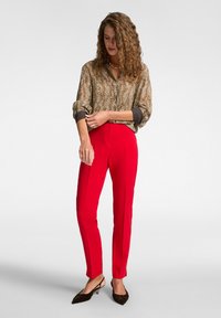Blusa con stampa animale marrone e beige, vestibilità larga con polsini scuri; pantaloni rossi su misura con cuciture frontali; scarpe marroni a punta.