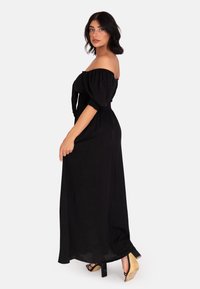 Abito lungo nero off-the-shoulder con maniche a sbuffo e corpetto arricciato. Presenta una gonna fluida, abbinata a tacchi alti neri.