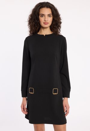 Femme avec des cheveux bouclés portant une robe noire à manches longues avec des boucles carrées dorées près de l'ourlet, se tenant devant un fond blanc uni.