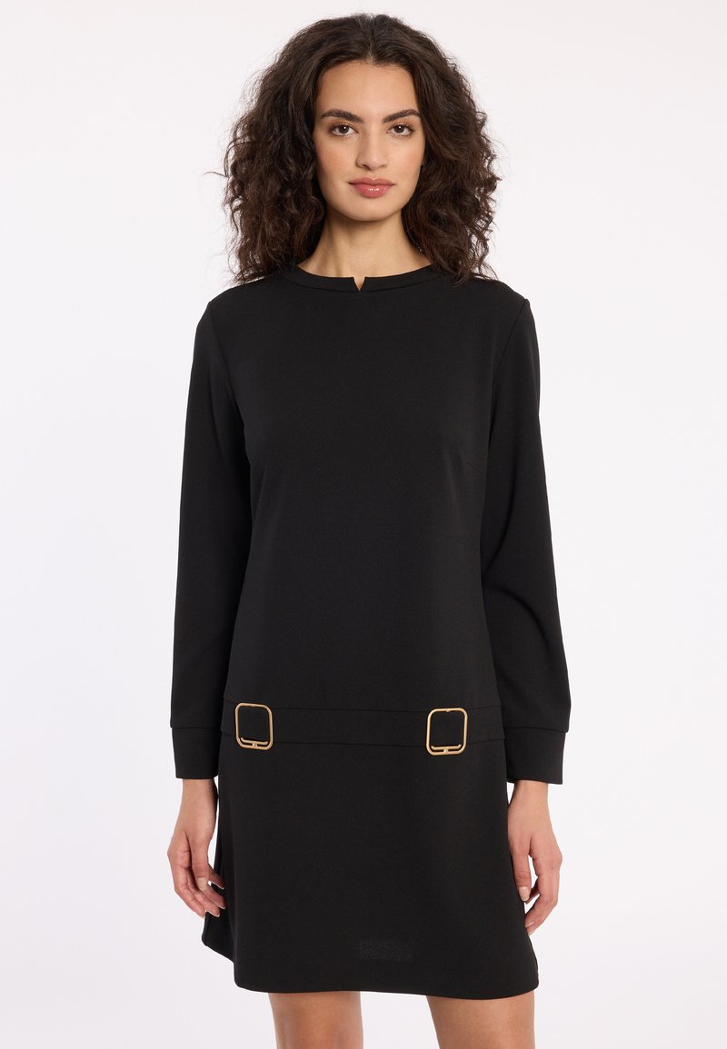 Woman with curly hair wearing a black long-sleeve dress with gold square buckles near the hem, standing against a plain white background.