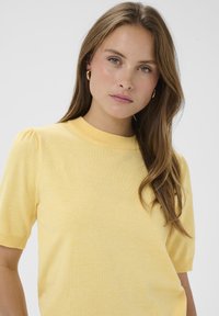 Hellgelbes Strickoberteil mit kurzen Ärmeln, hohem Ausschnitt und dezenten Puffärmeln. Glatte Textur und körpernahe Passform.