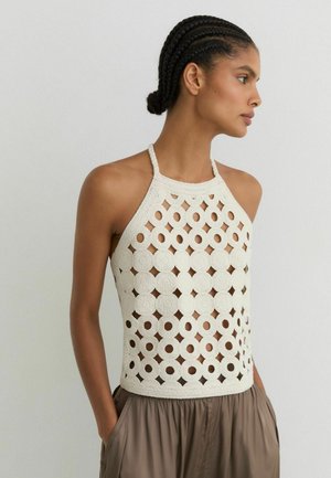 Žena s vrkočmi, oblečená v biely háčkovaný top bez rukávov a voľné hnedé nohavice, stojaca so založenými rukami v rukách pri bielej stene.