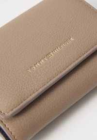 Beige nahkakotelo, jossa on teksturoitu pinta. Varustettu kultaisella kohokuvioidulla "TOMMY HILFIGER" -logolla ja erottuvalla ompeleella. Kompakti muotoilu.