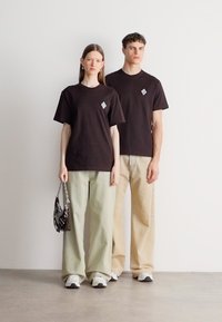 Czarne bawełniane t-shirty z diamentowym logo, zestawione z szerokimi spodniami w jasnym zielonym i beżowym kolorze. Dopełnieniem stylizacji są sneakersy.