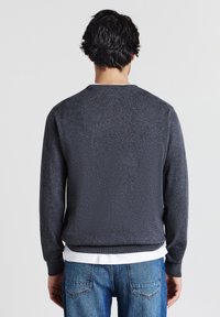 Mann, der mit dem Rücken zur Kamera steht, trägt einen dunkelgrauen Pullover über einem weißen Hemd und blauen Jeans vor einem einfarbigen Hintergrund.