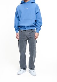 Person som bär en blå huvtröja med framficka, grå lösa jeans och vita sneakers, stående mot en vit bakgrund.