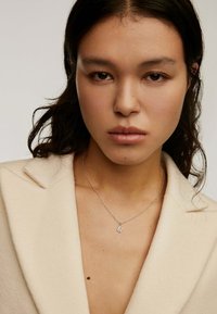 Collar de plata con un pequeño colgante en forma de "M", lucido sobre piel desnuda con un abrigo beige claro, entallado, que presenta solapas con muesca.