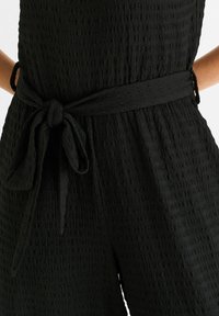Black textured jumpsuit with a tied waist belt. Material features a crinkled pattern. Sleeveless design, offering a relaxed fit around the hips.