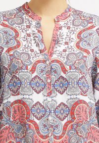 Weißes Hemd mit Knöpfen, das ein Paisleymuster in Rot, Blau und Pink zeigt, mit einem Mandarin-Kragen und Knopfverschlüssen. Glatte Stoffstruktur.