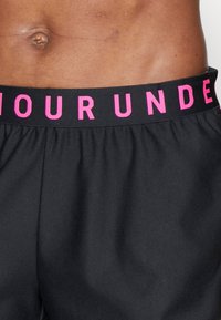 Närbild av en persons nedre torso iförd svarta shorts med svart midjeband som har rosa versaler.