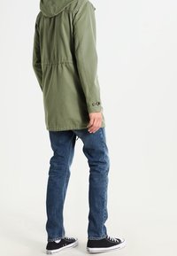 Olivově zelená kapucí vybavená bunda se stahovací šňůrkou, zapínáním na knoflíky na manžetách a rovný střihem, ve stylu s modrými džínami a černými teniskami.