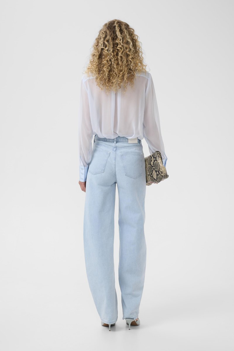 Vrouw met krullend blond haar staat met de rug naar de camera, draagt een doorschijnend wit blousje, lichtblauwe wijde jeans, witte hakken en houdt een clutch van slangenleer vast.