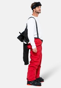 Pantalones de nieve rojos, camiseta blanca de manga larga, chaqueta de nieve negra y gafas de esquí. Tejido texturizado, tirantes ajustables y botas resistentes con detalles en naranja.