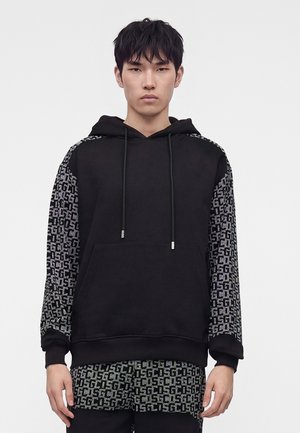 Jeune homme portant un sweat à capuche noir avec des manches à motifs et une chemise et un pantalon assortis à motifs sur fond uni.